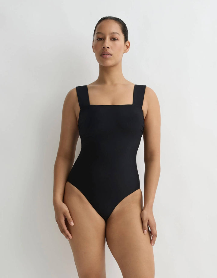 A woman modeling a solid black one-piece swimsuit with a square neckline and wide shoulder straps, standing against a plain light gray studio background.