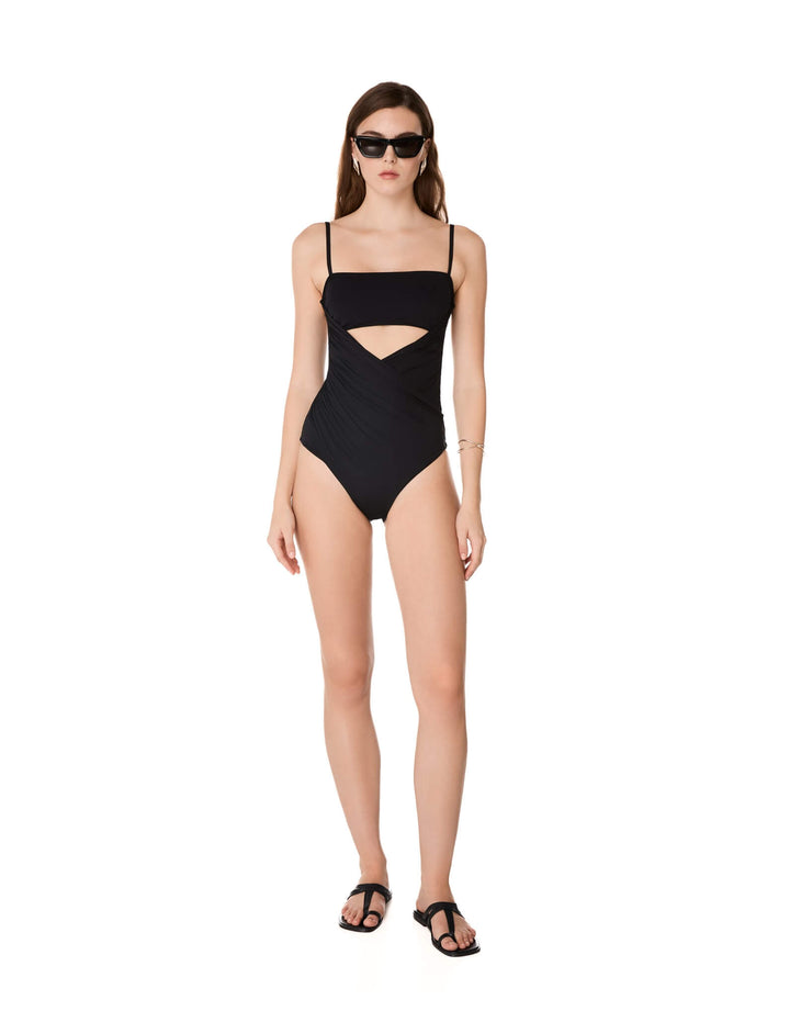 Kióhne Sierra black one-piece swimsuit with diagonal draped panels, bust cut-out and thin straps, styled with black sandals and sunglasses