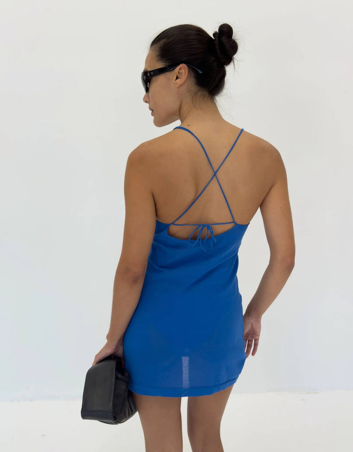 A woman shown from the back wearing a bright blue mini dress with thin crisscross back straps and holding a black clutch bag against a plain white studio background.